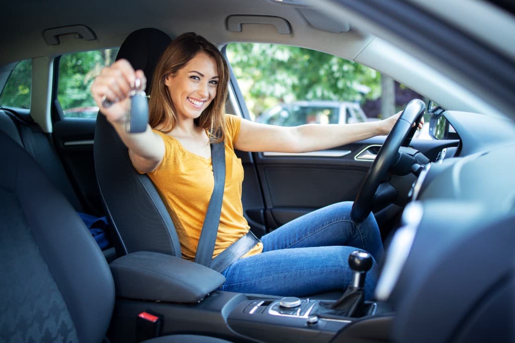 Aluna sorrindo ao volante, com o cinto afivelado e a chave do carro na mão.